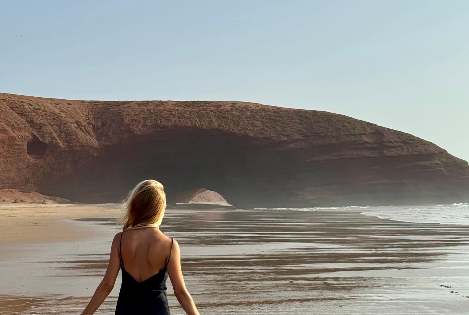 Maroc hors des sentiers battus : 1 semaine à Mirleft Legzira voaygeuse de dos au bord de la plage devant l'arche de pierre au coucher de soleil au maroc
